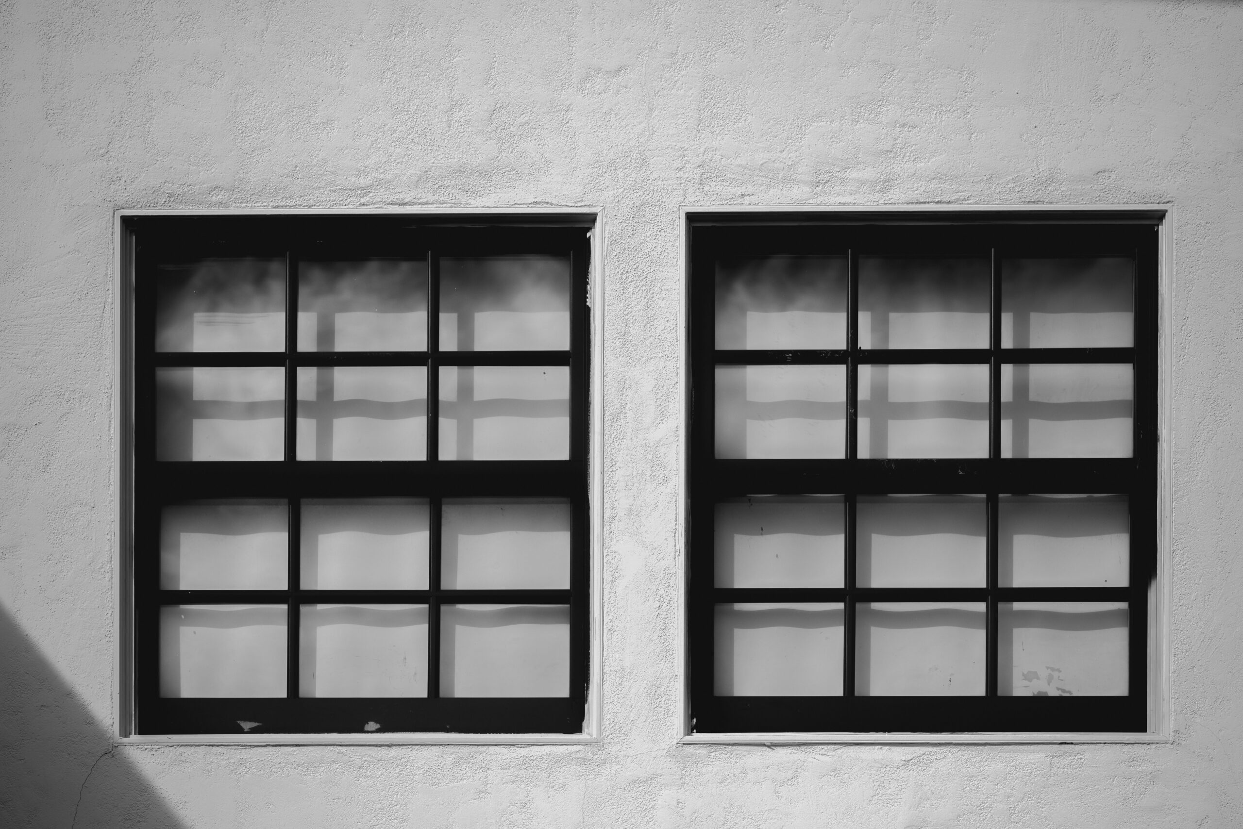 Due finestre a griglia nera su muro intonacato bianco, luce naturale riflessa sui vetri, stile minimalista architettonico